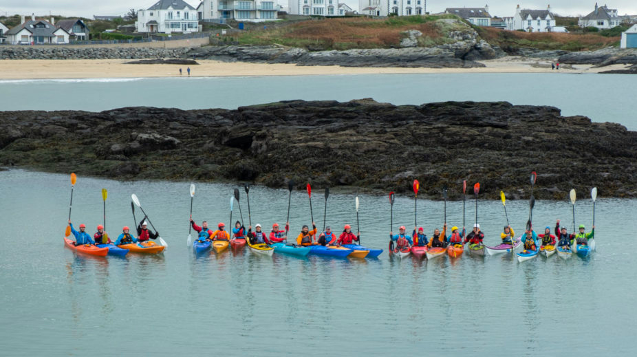 A colour photo of 22 sea kayakers in a line in their sea kayaks on the sea. They are lifting their paddles in the air. The kayakers are symbolising the number of people who are diagnosed with lobular breast cancer every day in the UK.