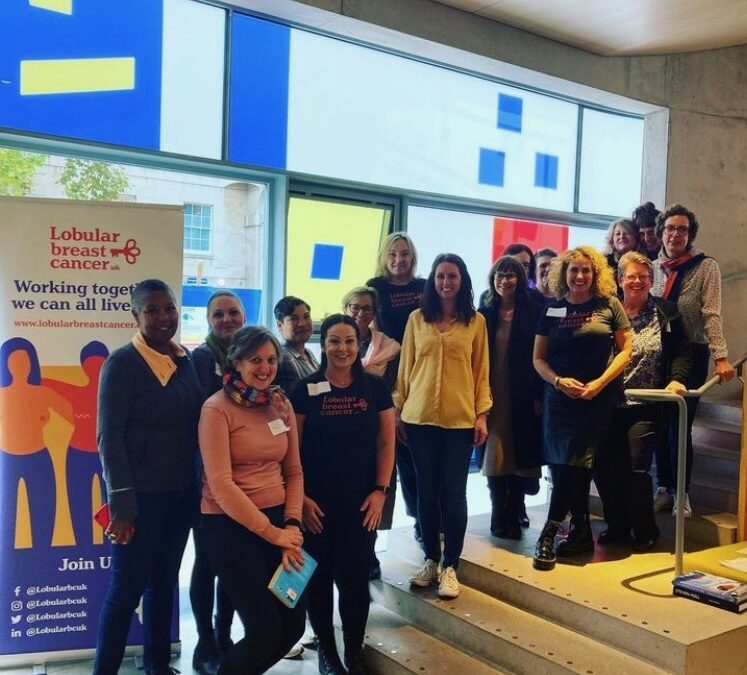 Colour photograph of 12 women standing looking to the camera next to a Lobular Breast Cancer UK banner. They have all taken part in LBCUK's first Wellbeing Workshop funded by the National Lottery Community Fund.