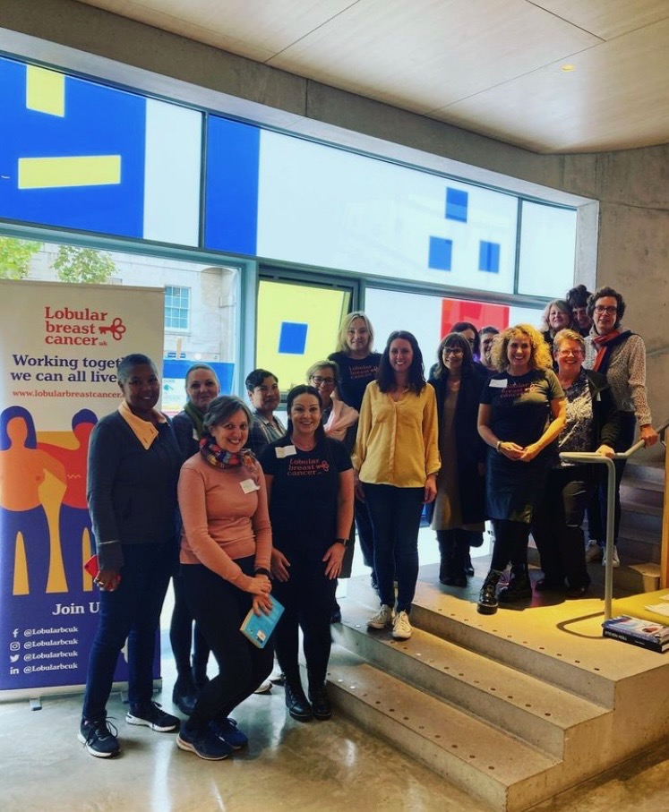 Colour photograph of 12 women standing looking to the camera next to a Lobular Breast Cancer UK banner. They have all taken part in LBCUK's first Wellbeing Workshop funded by the National Lottery Community Fund.