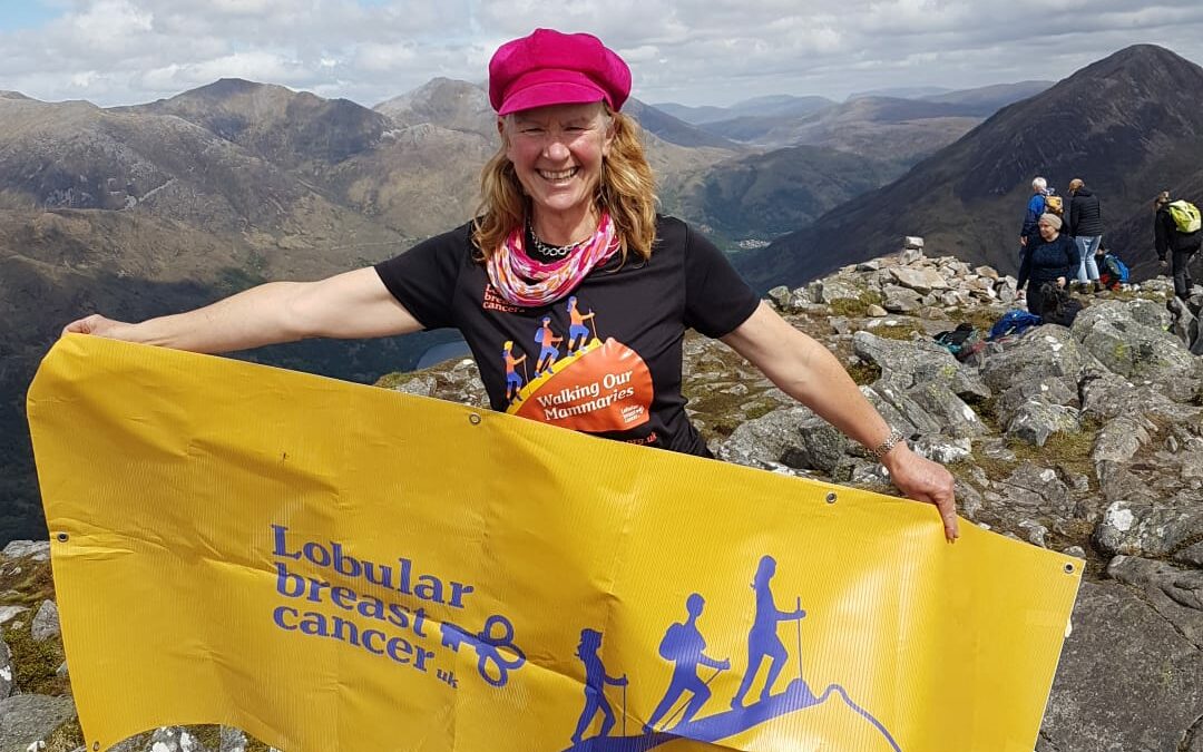 Colour photo of a woman standing on top of a mountain holding a yellow banner. In purple the banner states Lobular Breast Cancer UK and has a logo of women walking up a breast and the title Walking our Mammaries. The woman has a pink hat on and long red hair.