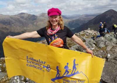 Colour photo of a woman standing on top of a mountain holding a yellow banner. In purple the banner states Lobular Breast Cancer UK and has a logo of women walking up a breast and the title Walking our Mammaries. The woman has a pink hat on and long red hair.