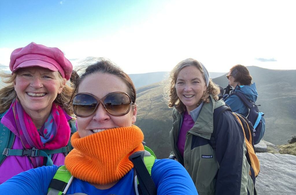 Four women standing on the top of the Moors on a cold but sunny day. Three are looking at the camera and smiling as they celebrate their summit of Kinder Scout, part of their training for a funder towards Lobular Breast Cancer. The fourth woman looks out over the views.