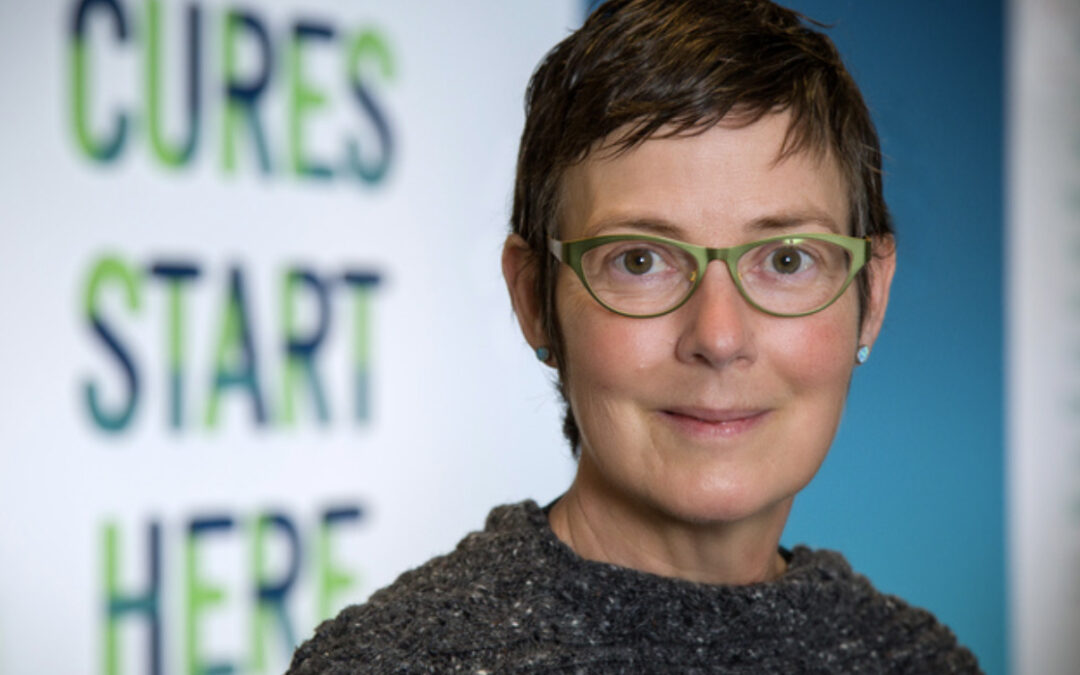Colour photograph of Lobular patient advocate Leigh Pate. Leigh has short dark hair and wears glasses. In the background in green, the words Cures Start Here are on the wall