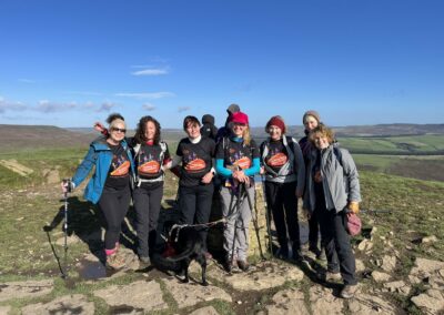 7 women walkers standing at a trig point wearing Lobular Breast Cancer Walking our Mammaries t-shirts